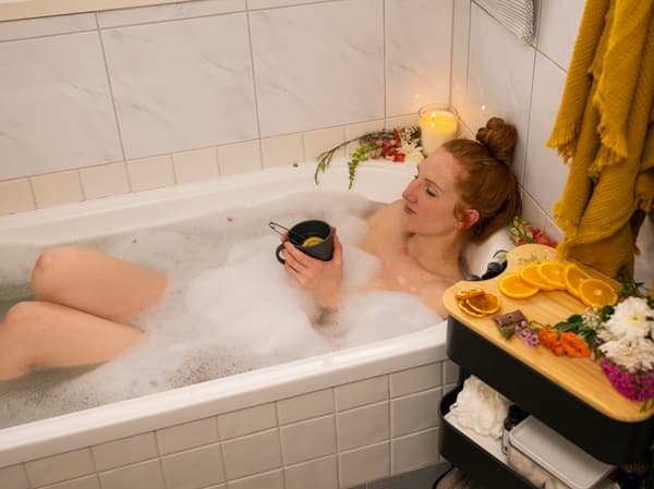 Een vrouw ligt in een bubbelbad met daarrond kaarsen. Ze heeft een warme kop thee in de hand en op het roltafeltje naast het bad liggen stukjes sinaasappel.