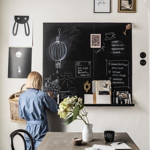 Een vrouw staat voor een krijtbord, ze is aan het krijten. Op het memobord staan verschillende boodschappen. Je ziet een huiselijke setting met een tafel, bloemen, een koffiekopje en wat accessoires op het krijtbord.