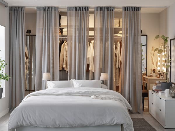 Een wit bedframe, een open kledingkast met daarvoor grijze gordijnen die de kast half bedekken, een witte ladekast en een grijs vloerkleed.