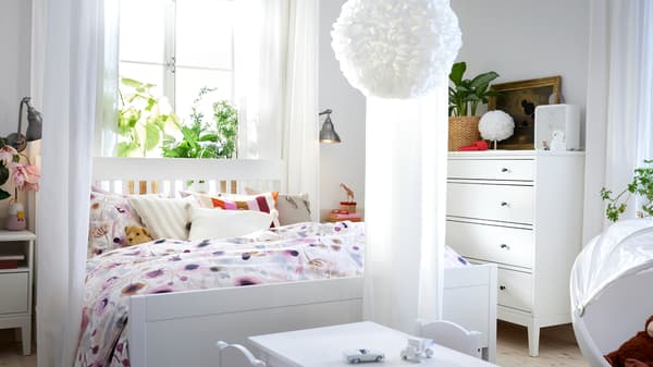 Een wit IDANÄS bed met LÖNNHÖSTMAL beddengoed staat naast een witte IDANÄS kast met vier lades voor een raam.