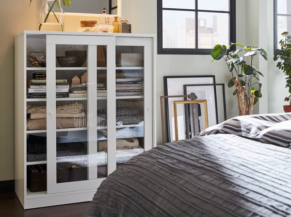 Een witte kast met glazen schuifdeuren, fotolijsten tegen de muur en een grijze sprei op het bed.