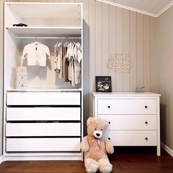 Een witte PAX kast met daarin babykleertjes, ernaast staat een witte ladekast en er voor staat een grote teddybeer 