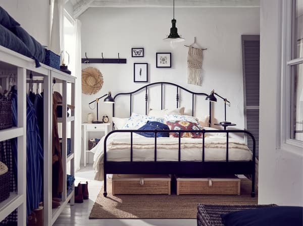 Een witte slaapkamer met een zwart stalen IKEA SAGSTUA bedframe, met een gewelfd hoofdeinde en messingkleurige details en opbergdozen.