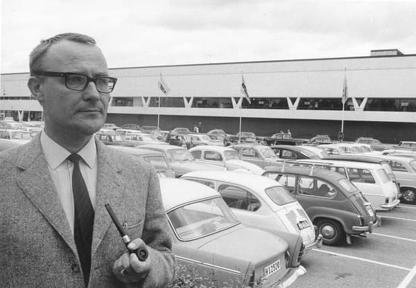 Een zwart-witfoto van de jonge Ingvar Kamprad die op de parkeerplaats van een IKEA-winkel staat en een pijp rookt.