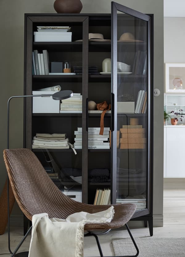 Een zwartbruine IKEA REGISSÖR vitrinekast vol doosjes, boeken en glazen, achter een fauteuil.