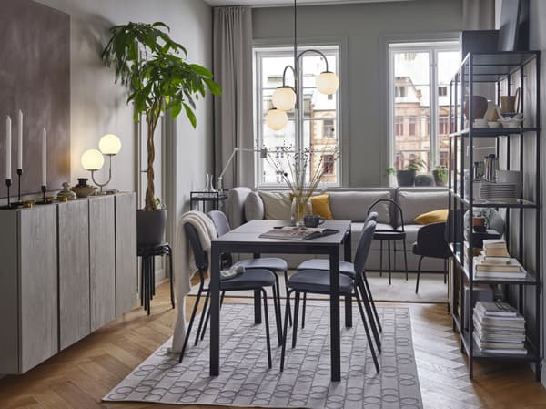 Een zwartbruine tafel en grijze stoelen staan op een tapijt met geometrisch patroon. Daarboven hangt een chromen hanglamp met 3 armen.