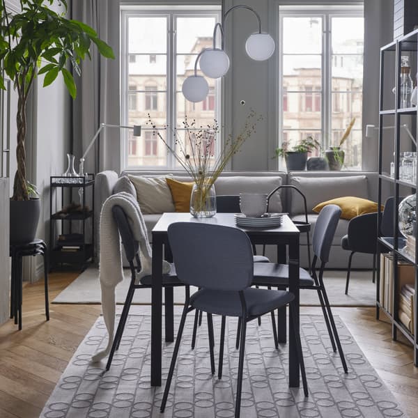 Een zwartbruine tafel, grijze stoelen, een tapijt met geometrisch patroon, een wit met chromen hanglamp en een lichtgrijze bank.
