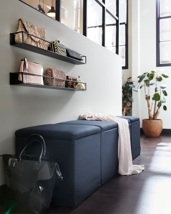 Een zwarte poef met opbergmogelijkheden, zwarte wandplanken met tassen erop en planten.