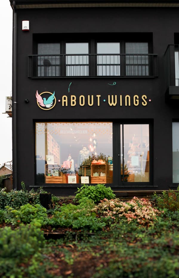 Een zwarte winkelgevel met het opschrift “ABOUT·WINGS”, een verlichte etalage en groene beplanting op de voorgrond.
