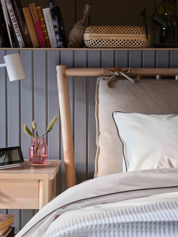Egy BJÖRKSNÄS ágy sarka látszik egy fa borítású fal előtt beépített könyvespolccal, az ágy mellett éjjeliszekrény látszik.