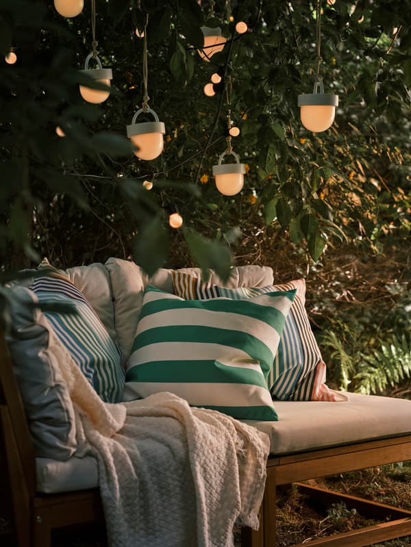 Egy fa mellett álló elemes NÄMMARÖ kanapé, rajta egy takaró és néhány díszpárna. Felette több SOMMARLÅNKE világítófüzér lóg.