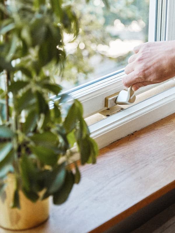 Egy fehér ablak fa párkányzsal, amelyen egy sárga cserépben lévő növény áll. Egy kéz fogja az ablakkilincset és nyitja az ablakot.
