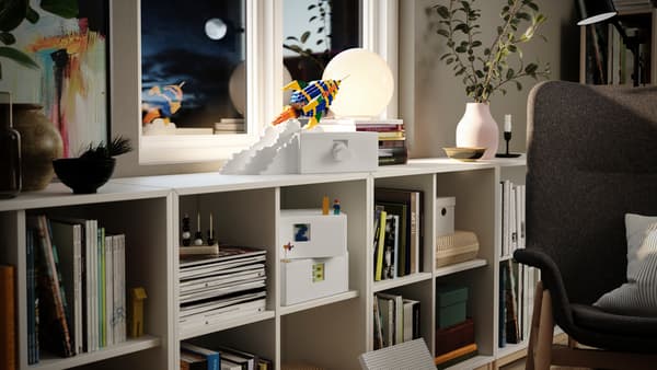 Egy fehér BYGGLEK doboz áll egy KALLAX tárolórendszerben, egy LEGO kockákból épített rakéta van a tetején.