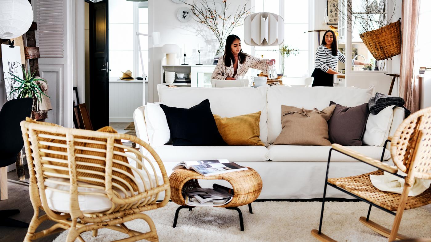 Egy fehér nappalisarokban két nő áll egy fehér BACKSÄLEN kanapé mögött, amely előtt szőnyeg, székek és lábtartó látható.