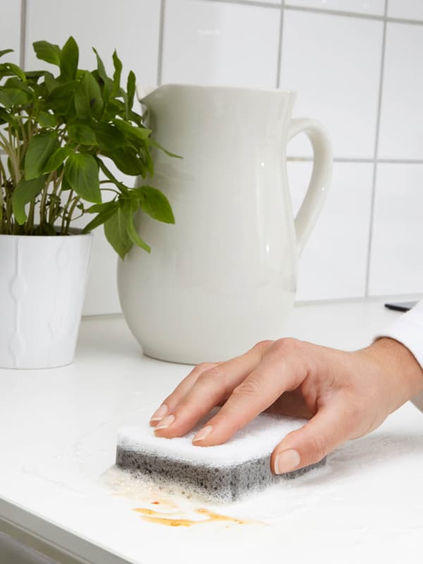 Egy fehér SVAMPIG szivacsot látunk, amint valaki éppen egy foltot töröl le vele egy fehér munkalapról. A háttérben egy fehér kancsó és egy fehér kaspóban zöld növény látható.