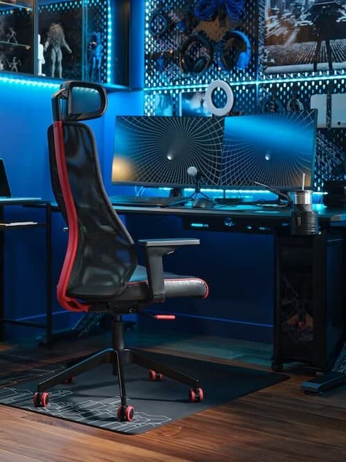 Egy fekete és piros MATCHSPEL gamer szék egy számítógépasztalnál. A monitor mögötti hirdetőtábla és a fali polcok kék LED-szalaggal vannak megvilágítva.