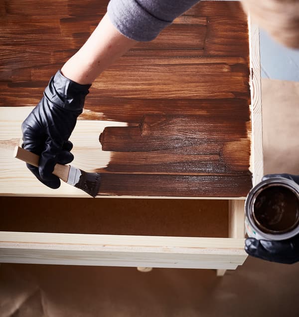 Egy fekete latex kesztyűt viselő lakberendező egy IKEA RAST 3 fiókos szekrényt sötétbarnára pácol. 