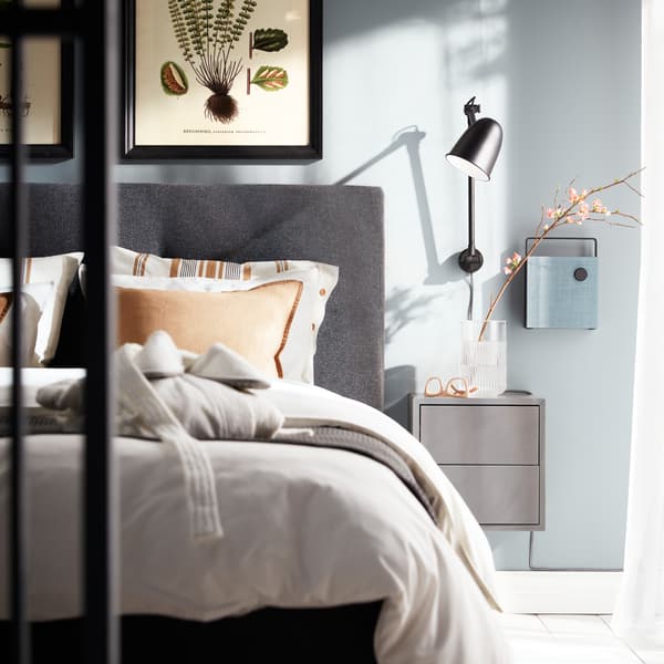 Egy fekete SKURUP falilámpa, egy sötétszürke EKET szekrény és egy falra szerelt Bluetooth hangszóró egy sötétszürke IDANÄS ágy mellett.