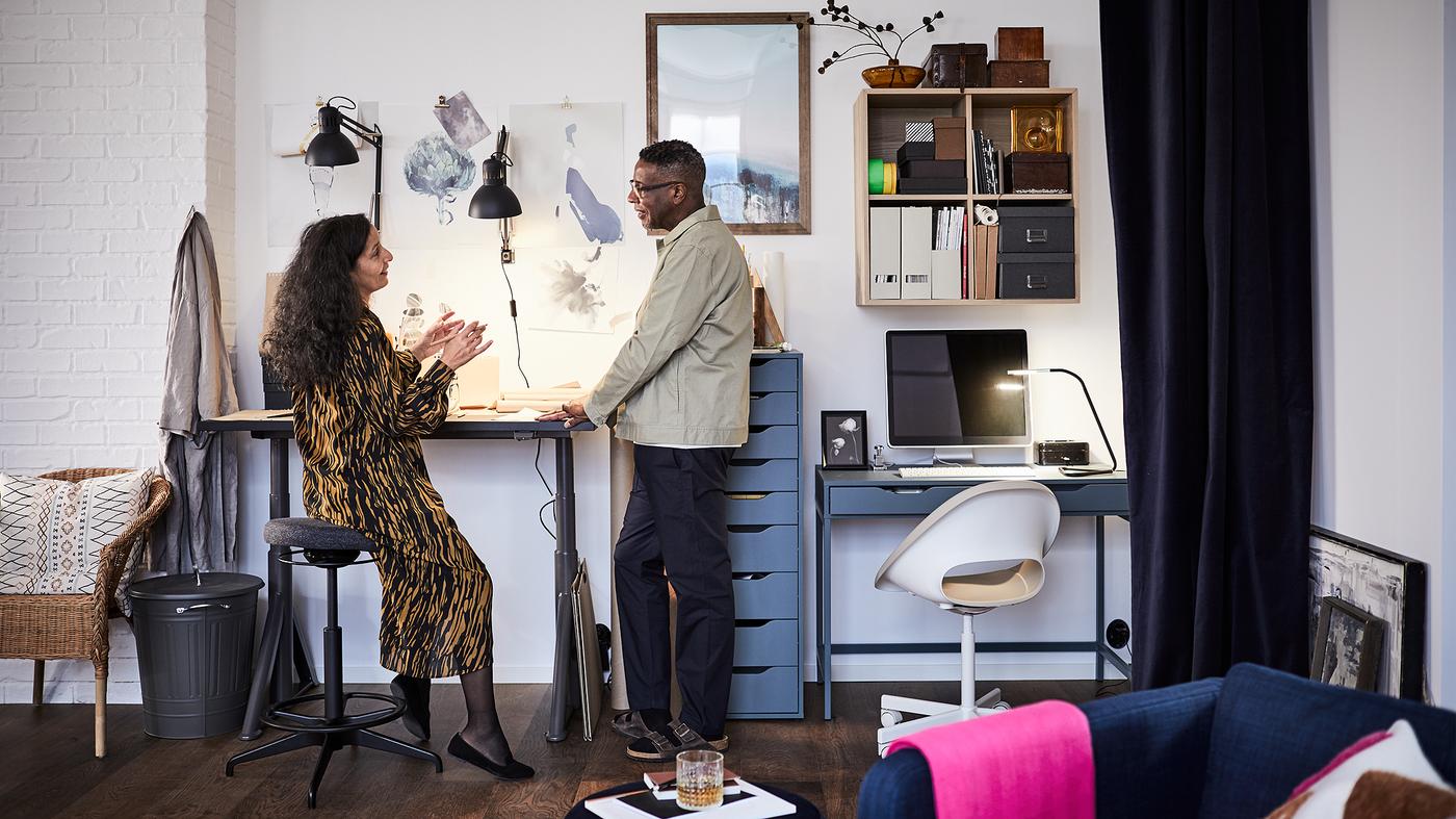 Egy férfi és egy nő egy otthoni irodában. A férfi egy IDÅSEN ülő/álló munkavégzéshez tervezett asztal mellett áll, a nő egy LIDKULLEN ülő-/állótámasznak dől.