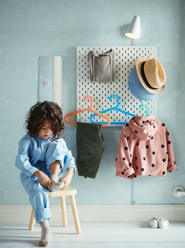 Egy gyerek zoknit húz egy ülőkén ülve. Mögötte a falon SKÅDIS lyukacsos tábla lóg, amire különféle ruhadarabok vannak felakasztva.