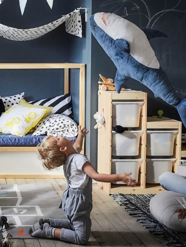 Egy gyerekszobában a HEMMAHOS szőnyegen térdelő gyereket látunk, amint a BLÄHAJ puha játék cápát dobálja a magasba.