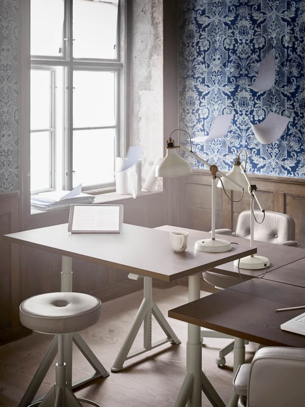 Egy irodában négy, állítható magasságú IDÅSEN íróasztalt látunk. Az iroda falait fehér virágmintás kék tapéta borítja.