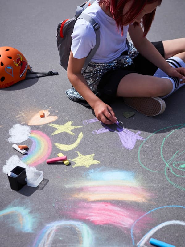 Egy járdán ülő lány, amint éppen MÅLA krétával rajzol az aszfaltra.