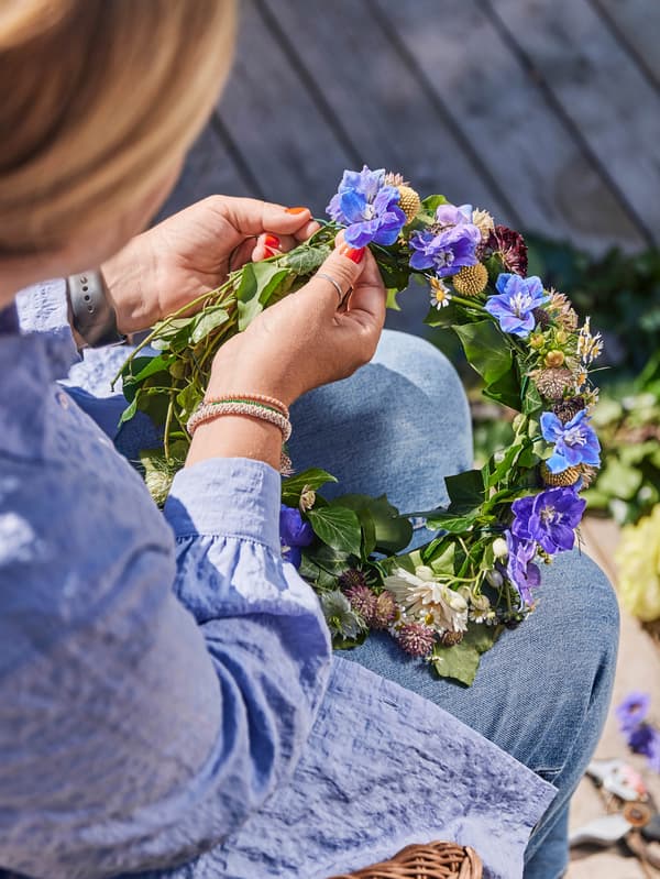 Egy kék ruhás nő Midsommar alkalmából virágkoszorút fon.