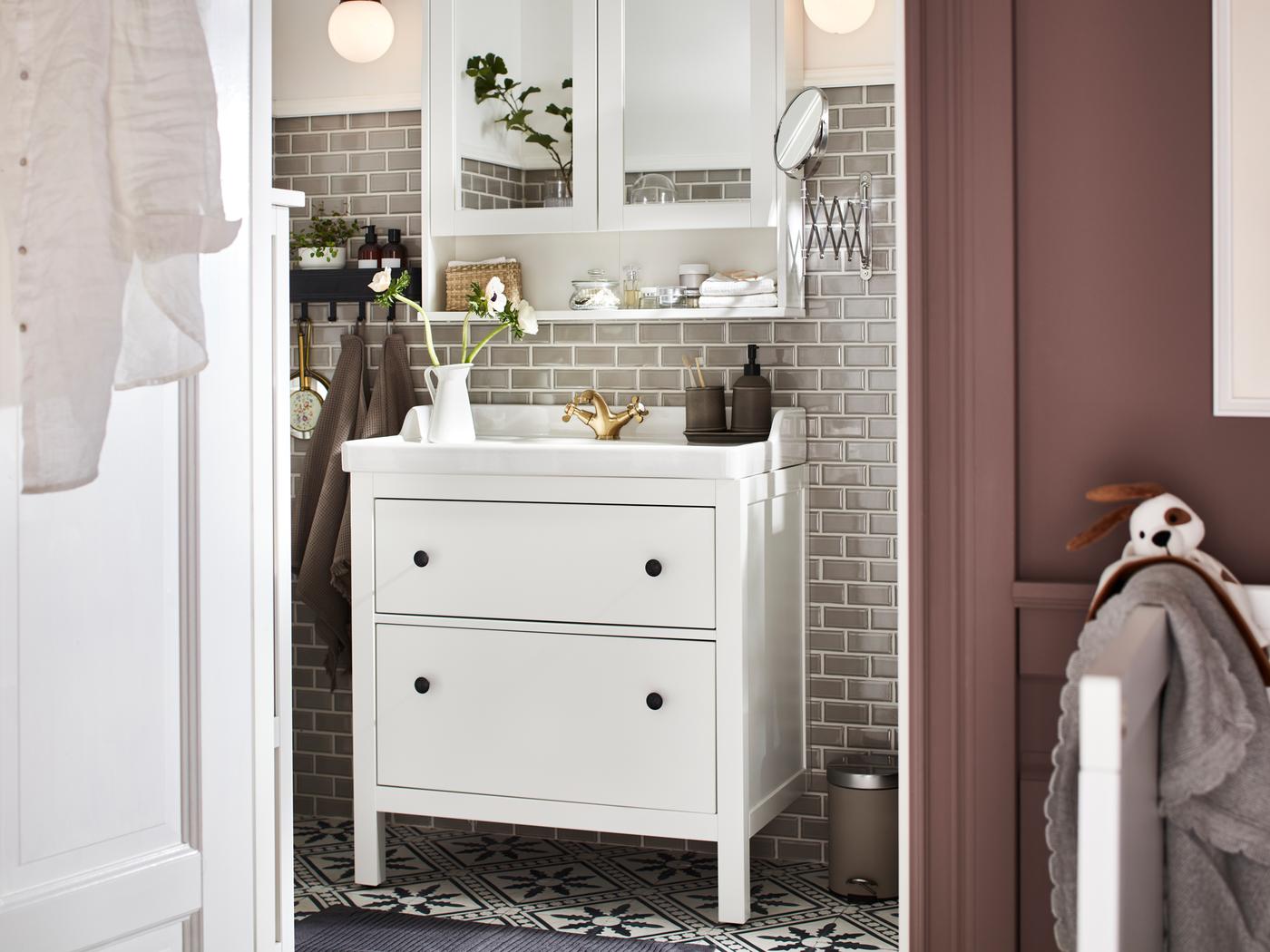 Egy kétfiókos HEMNES/RÄTTVIKEN mosdóállvány és egy fehér színű, kétajtós HEMNES tükrös szekrény egy fürdőszobában.