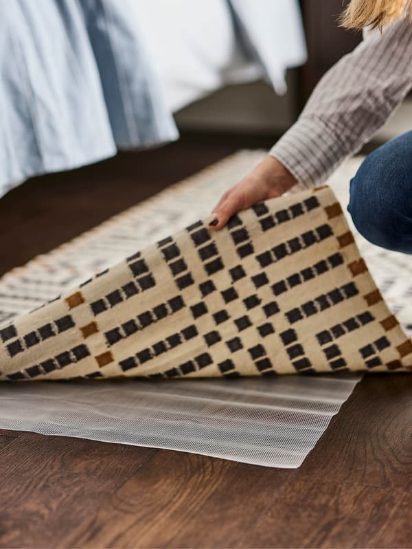 Egy kéz, amint felemeli a VAMDRUP szőnyeg sarkát. A szőnyeg alatt jól látszik a STOPP csúszásgátló alátét.