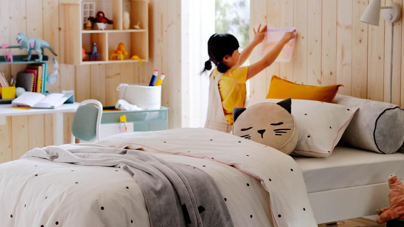 Egy kislány, amint éppen egy festményt tűz a világos színű, fából készült bútorokkal berendezett szoba falára. A háttérben egy RELATERA asztal ülő-/álló munkához áll, rajta különböző kiegészítőkkel.