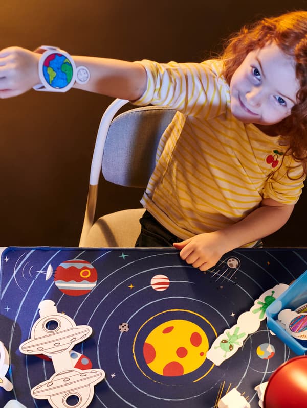 Egy kislány egy kivágott és kiszínezett AFTONSPARV papírkarórát mutat egy AFTONSPARV asztali alátét fölött.