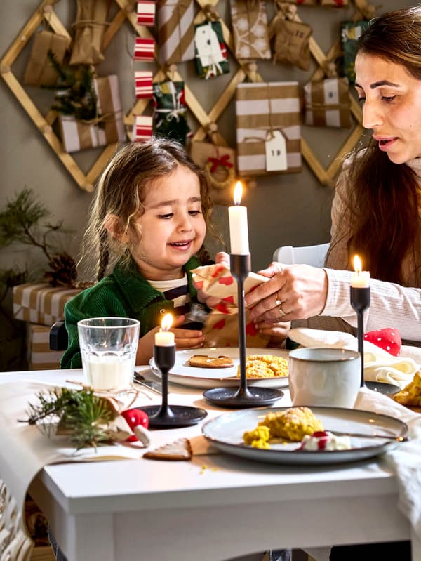 Egy kislány és édesanyja látható egy GLADELIG étkészlettel megterített asztal mellett. Az asztal közepén fekete FULLTALIG gyertyatartókban JUBLA gyertyák égnek.