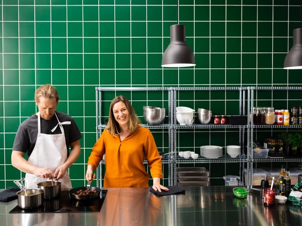Egy konyhában, melyet a rozsdamentes acél és zöld csempe dominál, egy nő és férfi áll, húsmentes golyókkal teli serpenyővel a kezükben.