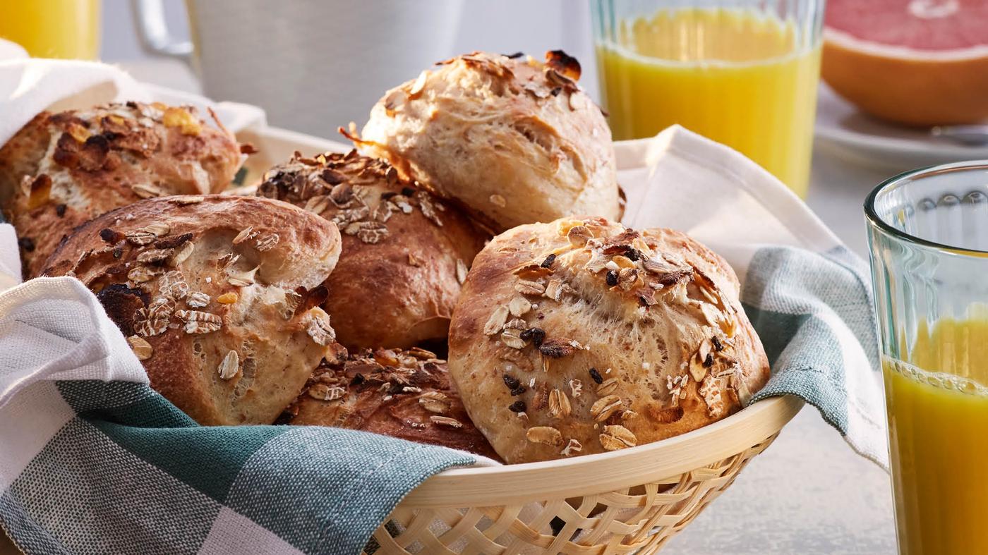 Egy kosár frissen sült, hidegen kelesztett zsemle müzlivel és sópehellyel, konyharuhán, kávé, tea, narancslé és grapefruit társaságában.