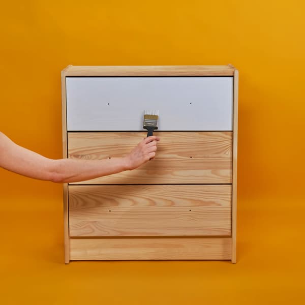 Egy lakberendező ecsetet tart, mellyel fehérre festi a RAST komód fiókjait.