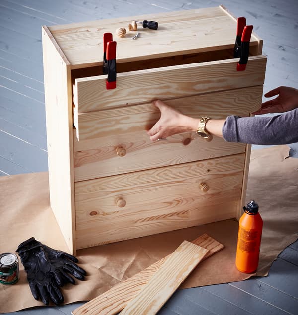 Egy lakberendező miközben felújít egy 3 fiókos IKEA RAST komódot a felső fiók elülső részét szorítókkal rögzíti.  