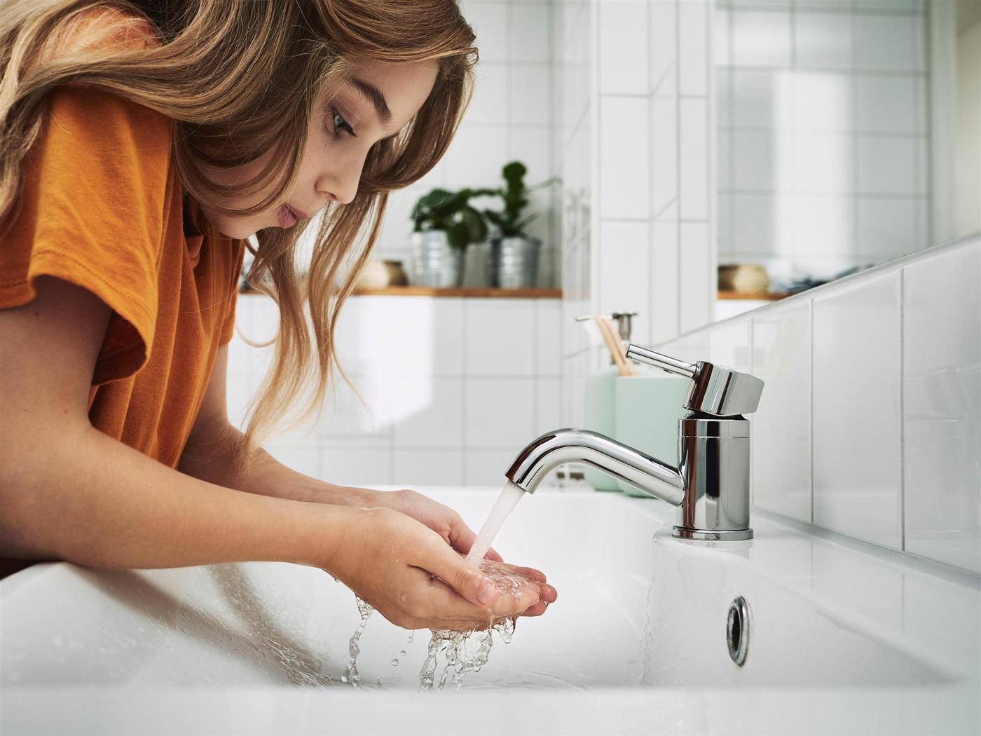 Egy lány egy fehér csempés fürdőszobában vizet folyat a csapból a mosdóba