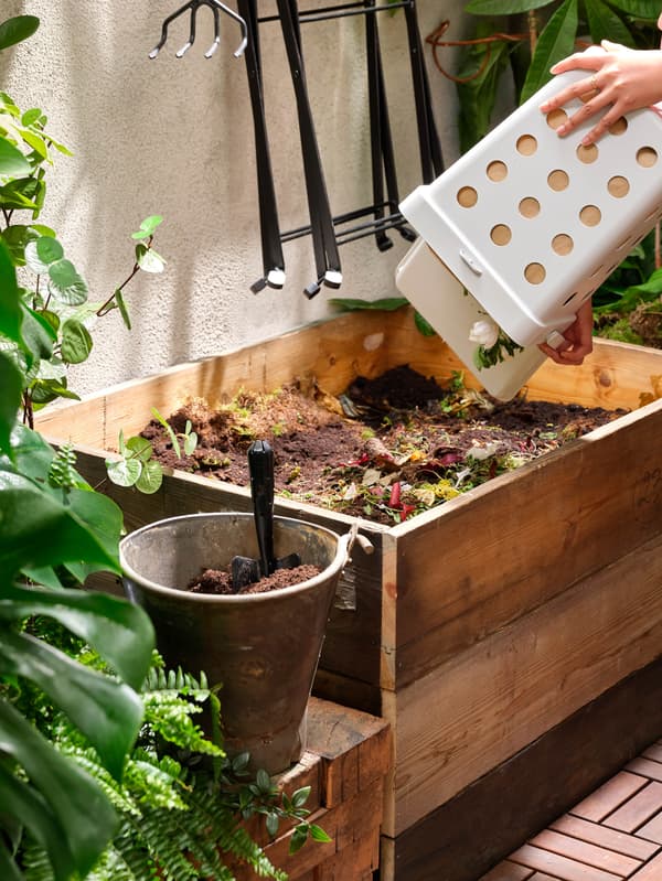 Egy nő egy fehér HÅLLBAR élelmiszerhulladék-tartót ürít ki egy deszkákból készített komposztálóba, amit apró, sok cserepes növénnyel díszített udvarban helyeztek el.