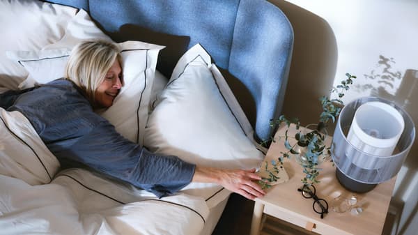 Egy nő fekszik egy kék kárpitozott ágyban, fehér lepedővel, és elnyújtózik egy fa éjjeliszekrényen. Az asztalon egy növény és egy hangszórós lámpa áll.