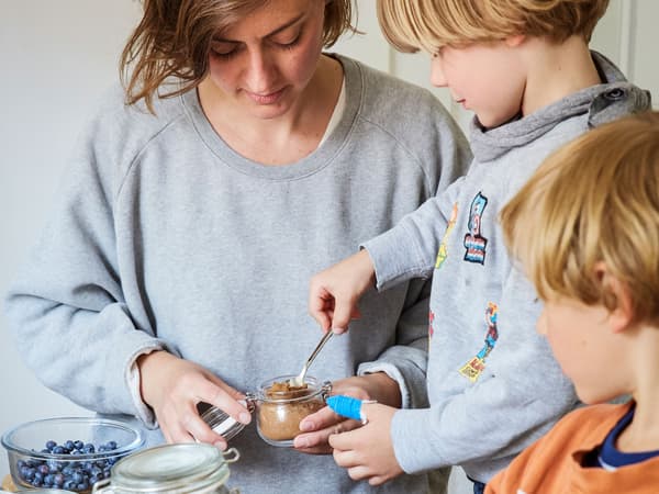 Egy nő két gyermekével reggelit készít csatos üveg edényben, melyben joghurt és granola van.