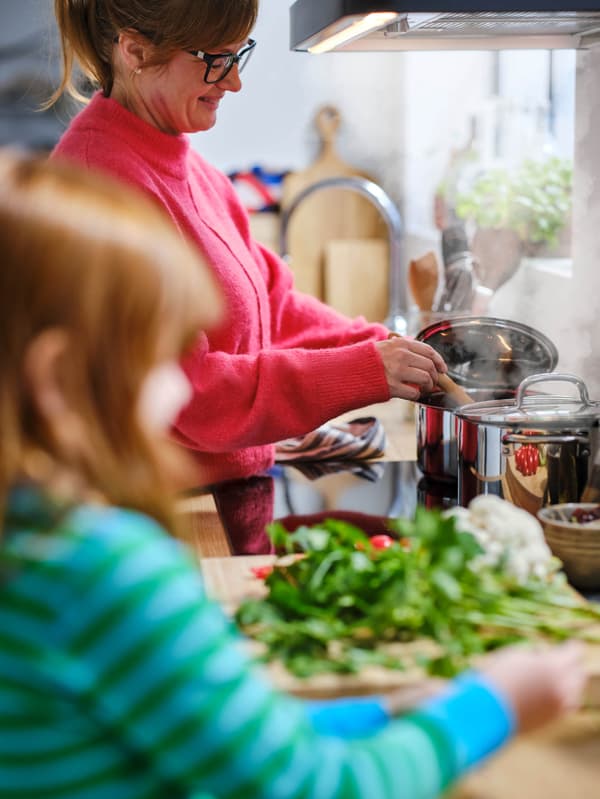 Egy rózsaszín pulóvert viselő nő két IKEA 365+ edényben főzi az ételt. Az előtérben egy kislány kék-zöld csíkos pulóverben friss fűszernövényeket aprít egy vágódeszkán.