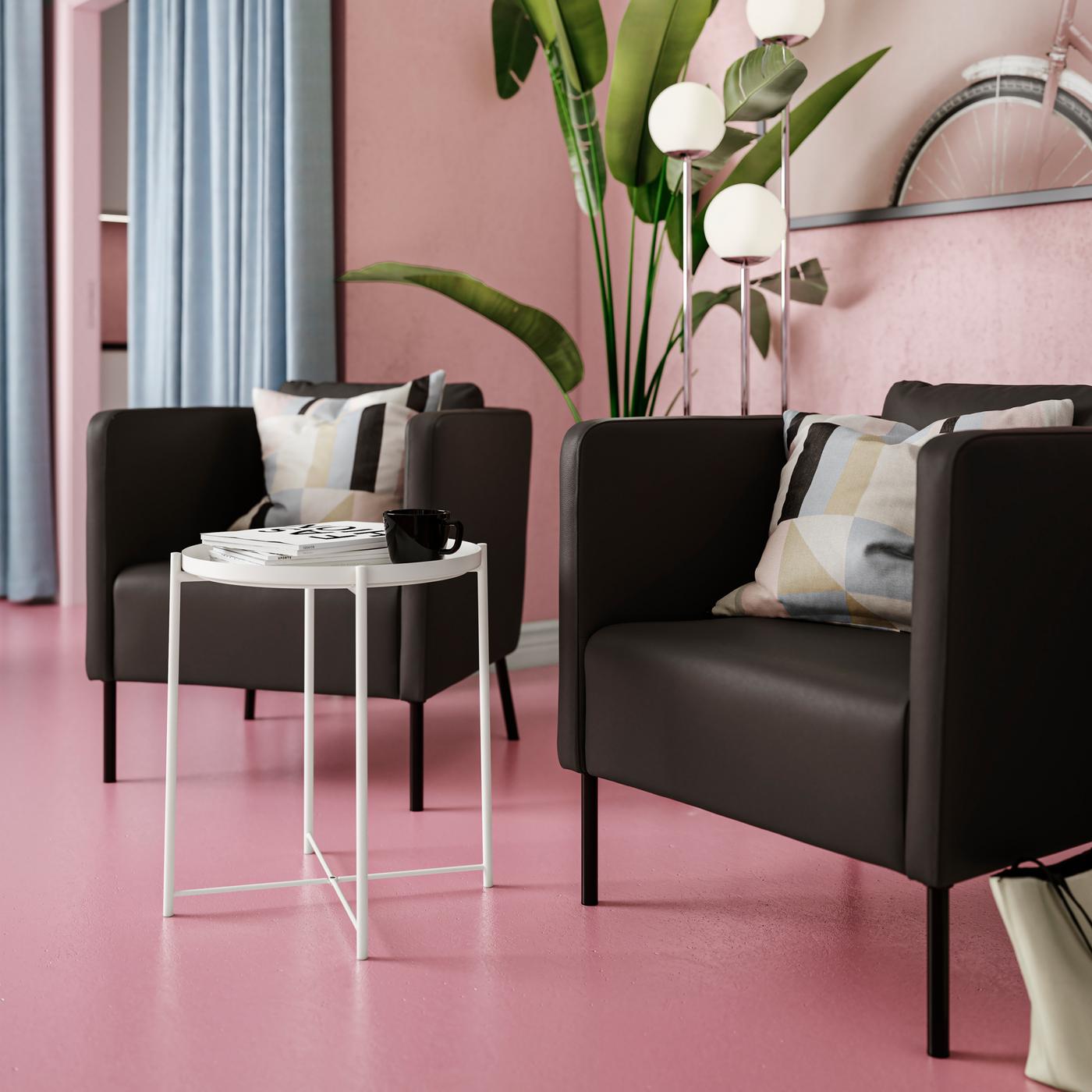 Egy rózsaszín szobában kerek tálca-asztal körül két szögletes formájú, modern stílusú EKERÖ fotel áll fekete bőrt imitáló huzattal. 