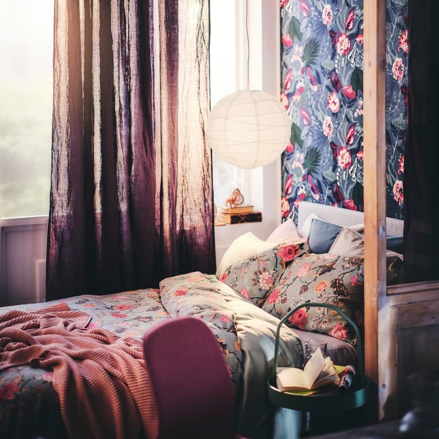 Egy sötét, virágos falon melletti ágy mindkét oldalán fekete SKURUP álló-/olvasólámpák vannak, az ablakban pedig egy TÄRNABY asztali lámpa.