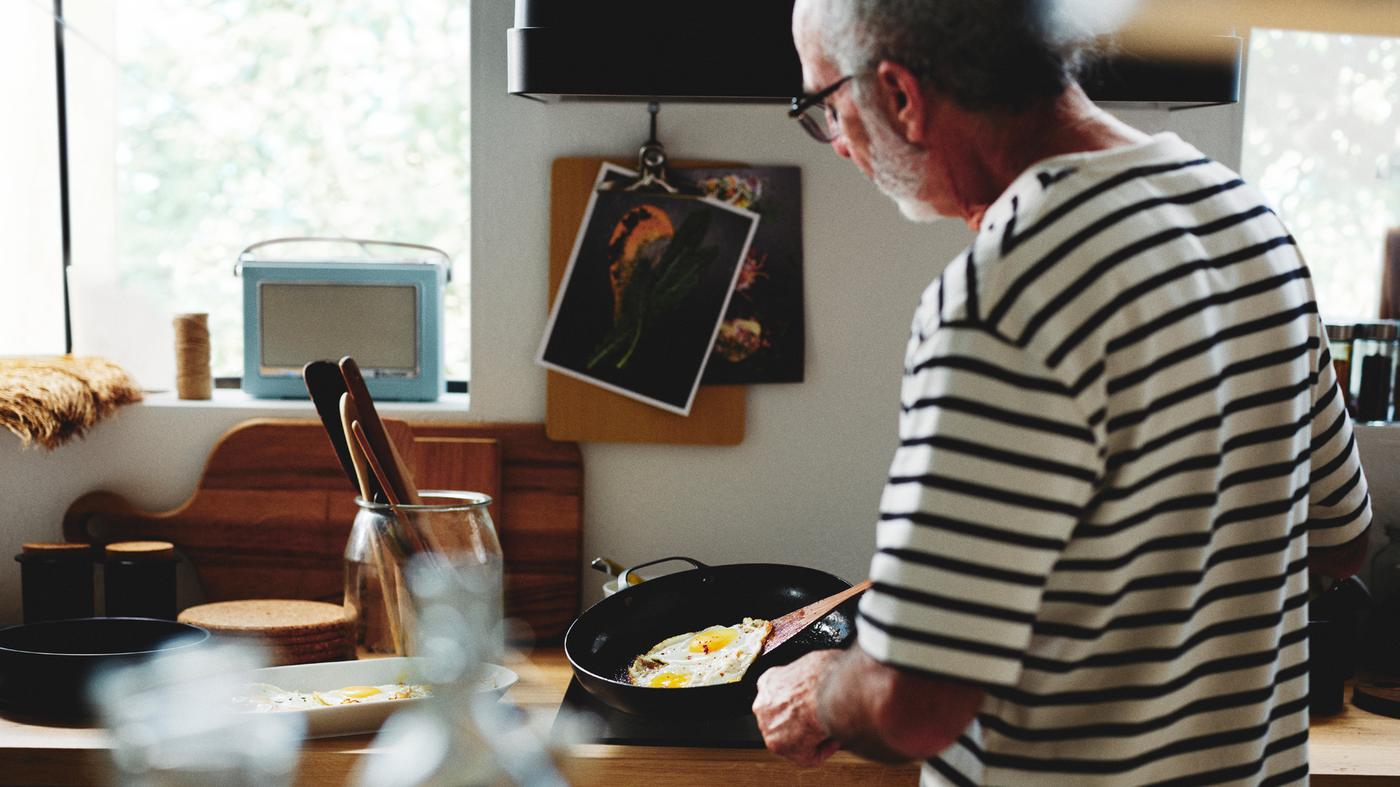 Egy személy áll egy konyhában, és egy szénacél VARDAGEN serpenyőben tojást süt. Mellette, egy másik VARDAGEN edényben konyhai eszközök láthatók.