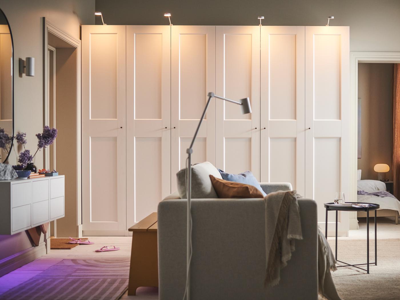 Egy szoba középen egy 2-személyes, bézs VIMLE kanapé, a hátsó fal mellett pedig két fehér PAX/GRIMO gardróbszekrény áll.