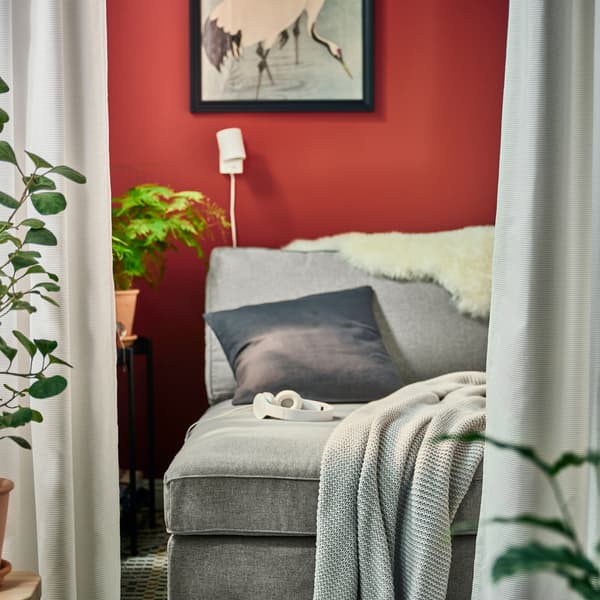 Egy szürke KIVIK fekvőfotel, rajta egy világosszürke INGABRITTA takaró és egy antracitszínű díszpárna, valamint egy fehér NYMÅNE falilámpa.