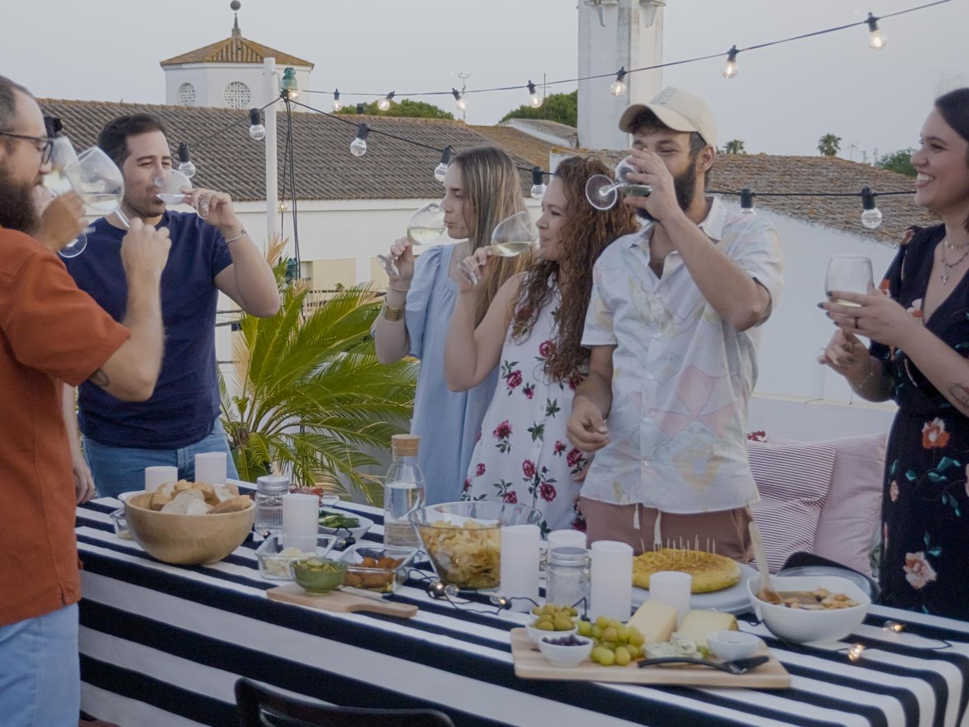 Egy teraszon a szabadban, étellel gazdagon megrakott büféasztalnál álló fiatal baráti társaság vidáman eszik, iszik és beszélget.