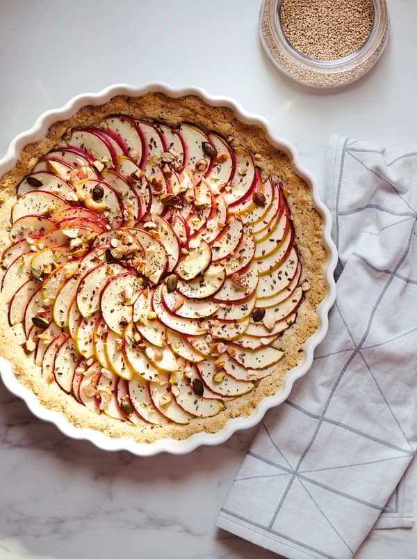 Egy torta, vékony almaszelettel, magvakkal és diófélékkel borítva egy fehér VARDAGEN pite edényben, márvány konyhai munkalapon állva.