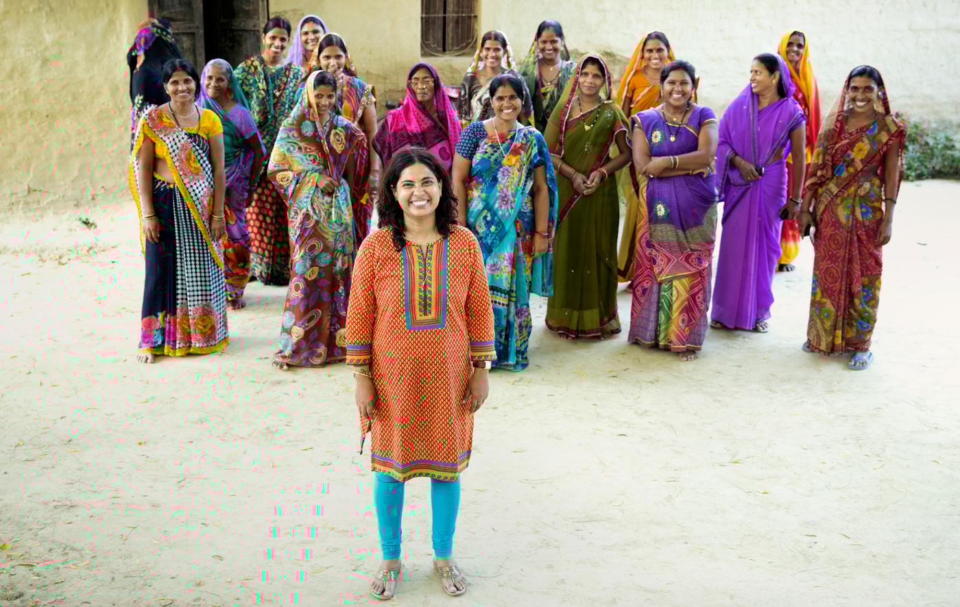 Ei gruppe med rundt 15 kvinner i lange, fargerike kjoler står smilende på en grusvei i en by.